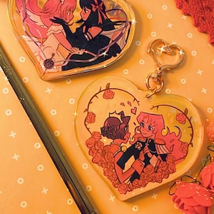 Op de afbeelding: Twee hartvormige sleutelhangers met illustraties van personages in anime-stijl. Eén sleutelhanger heeft twee figuren en de andere heeft twee figuren omringd door rozen. Beide hebben goudkleurige hartvormige sluitingen. Er is ook een decoratief zwaard aanwezig.