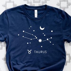 May include: Navy blue t-shirt featuring a white Taurus constellation design with a crescent moon and stars. The word "TAURUS" is printed below the constellation. The shirt is made of a soft material.