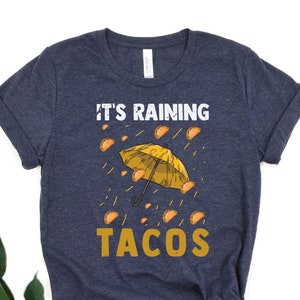 Peut inclure: Un t-shirt bleu marine avec un imprim&eacute; blanc d'un parapluie jaune avec des tacos qui tombent du ciel. Le texte "It's raining tacos" est imprim&eacute; sous le parapluie.
