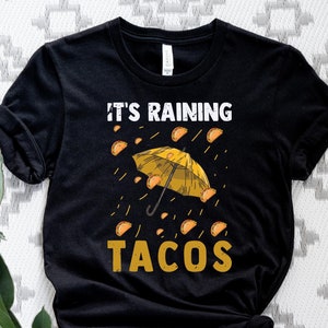 Peut inclure: T-shirt noir avec un graphique de tacos jaunes qui pleuvent du ciel avec un parapluie jaune. Le texte "It's Raining Tacos" est imprim&eacute; sur le t-shirt.