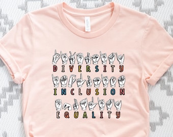 May include: A light pink t-shirt with the words "DIVERSITY, INCLUSION, EQUALITY" spelled out in colorful American Sign Language hand gestures. The shirt is made of soft material and has short sleeves.