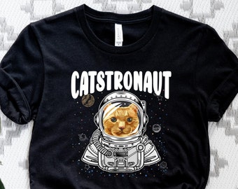 May include: Black t-shirt with a white graphic of a cat wearing an astronaut helmet. The text "CATSTRONAUT" is printed in white above the cat.