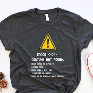 May include: A dark gray t-shirt with a white graphic of a warning sign and the text "ERROR <404>: COSTUME NOT FOUND."  The text below the graphic reads "PRESS ENTER TO RETURN TO OCTOBER 30TH. PRESS CTRL + ALT + DEL TO RESTART HALLOWEEN PRESS F6 TO GENERATE GHOST COSTUME."