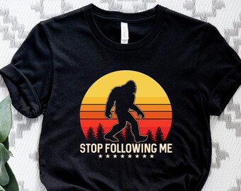 May include: Black t-shirt with a retro-style graphic of a Bigfoot walking in front of a sunset. The text "STOP FOLLOWING ME" is printed below the graphic.