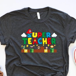 Puede incluir: Camiseta gris oscuro con un gr&aacute;fico de un personaje de videojuego y el texto "SUPER TEACHER".