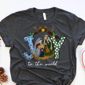 May include: A dark gray t-shirt with a Christmas-themed design featuring the word "JOY" with a nativity scene inside the letters. The text "to the world" is printed below the design.