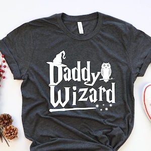 May include: A dark grey t-shirt with a white graphic that says "Daddy Wizard" with a wizard hat and owl illustration.