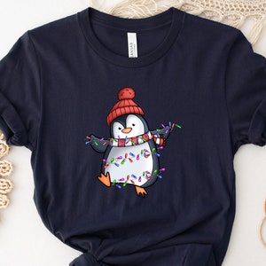 May include: A navy blue t-shirt with a cartoon penguin wearing a red Santa hat and a scarf, holding colorful Christmas lights.