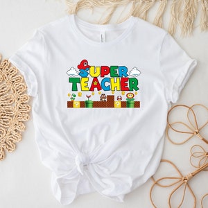 Puede incluir: Camiseta blanca con un gr&aacute;fico colorido de un dise&ntilde;o tem&aacute;tico de Super Mario Bros. que dice "SUPER TEACHER".