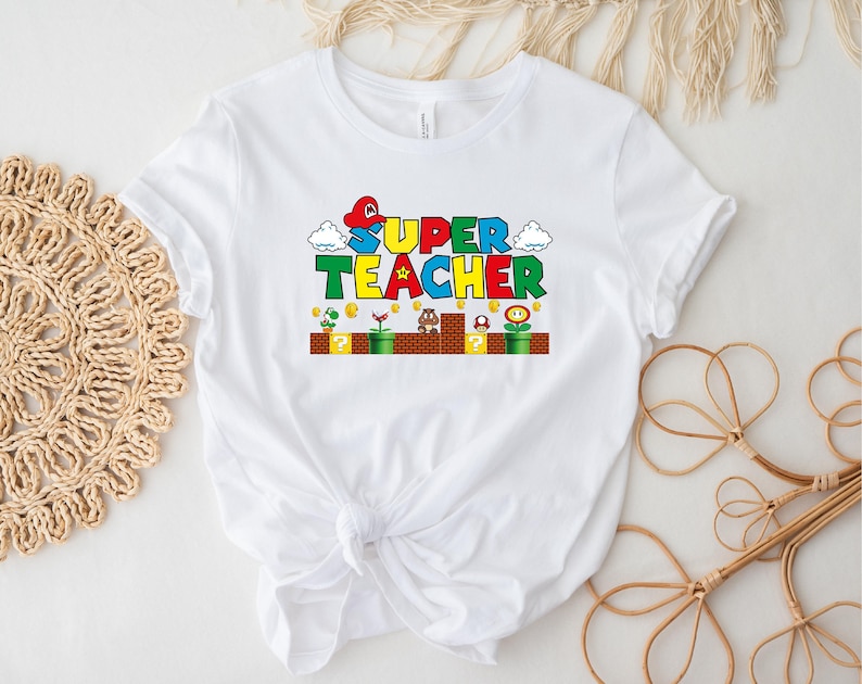 Puede incluir: Camiseta blanca con un gr&aacute;fico colorido de un dise&ntilde;o tem&aacute;tico de Super Mario Bros. que dice "SUPER TEACHER".