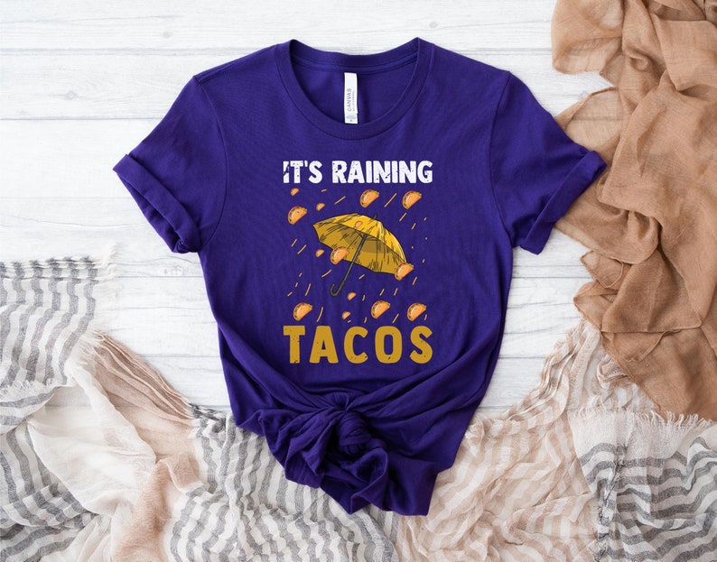 Peut inclure: T-shirt violet avec un parapluie jaune et le texte "It's Raining Tacos". Des tacos jaunes tombent du parapluie.