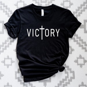 K&ouml;nnte beinhalten: Schwarzes T-Shirt mit einem wei&szlig;en Kreuz und dem Wort "VICTORY" auf der Vorderseite.