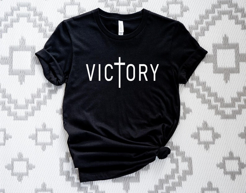 K&ouml;nnte beinhalten: Schwarzes T-Shirt mit einem wei&szlig;en Kreuz und dem Wort "VICTORY" auf der Vorderseite.