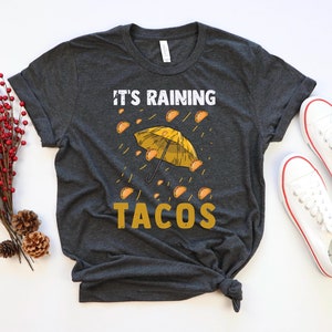 Peut inclure: Un t-shirt gris fonc&eacute; avec le texte "It's raining tacos" en lettres jaunes. Un parapluie jaune avec des tacos tombant du ciel est repr&eacute;sent&eacute; au-dessus du texte.