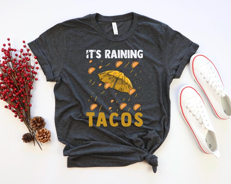 Peut inclure: Un t-shirt gris fonc&eacute; avec le texte "It's raining tacos" en lettres jaunes. Un parapluie jaune avec des tacos tombant du ciel est repr&eacute;sent&eacute; au-dessus du texte.