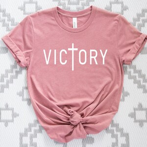 K&ouml;nnte beinhalten: Ein rosa T-Shirt mit einem wei&szlig;en Kreuz und dem Wort "VICTORY" auf der Vorderseite.