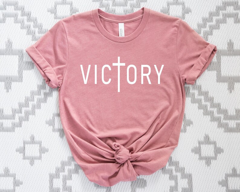 K&ouml;nnte beinhalten: Ein rosa T-Shirt mit einem wei&szlig;en Kreuz und dem Wort "VICTORY" auf der Vorderseite.