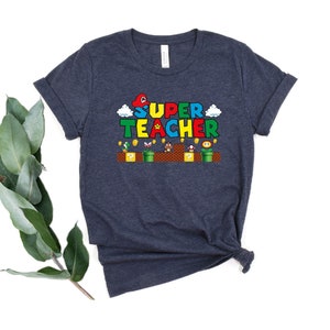 Puede incluir: Una camiseta azul oscuro con un dise&ntilde;o tem&aacute;tico de Super Mario Bros. El texto "SUPER TEACHER" est&aacute; en una fuente pixelada de colores.