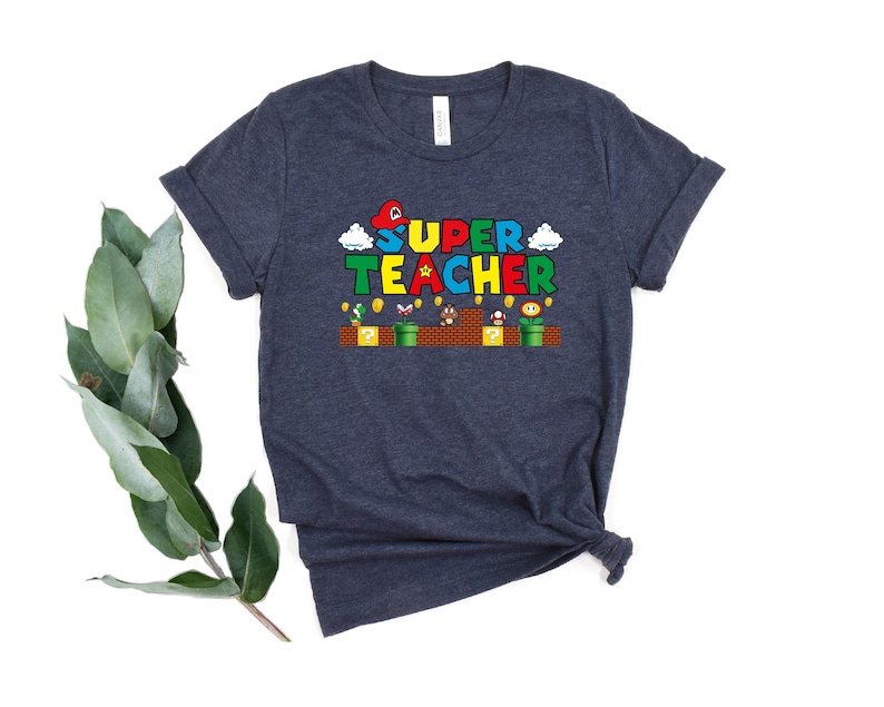 Puede incluir: Una camiseta azul oscuro con un dise&ntilde;o tem&aacute;tico de Super Mario Bros. El texto "SUPER TEACHER" est&aacute; en una fuente pixelada de colores.
