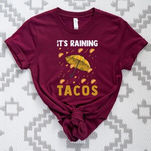 Peut inclure: Un t-shirt marron avec le texte "Il pleut des tacos" et un parapluie jaune avec des tacos qui tombent du ciel.