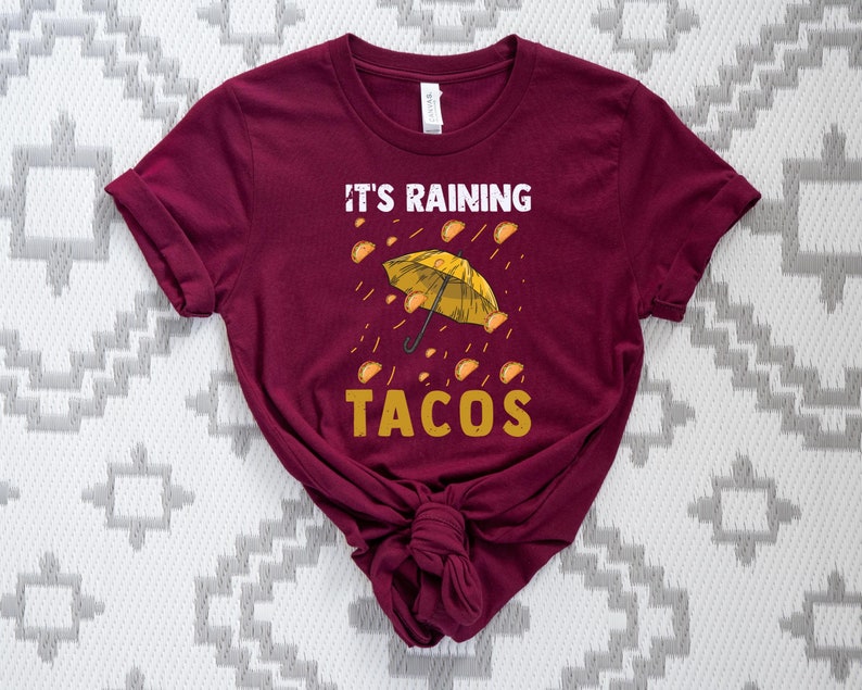 Peut inclure: Un t-shirt marron avec le texte "Il pleut des tacos" et un parapluie jaune avec des tacos qui tombent du ciel.