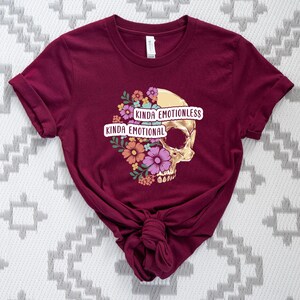Puede incluir: Una camiseta color burdeos con un gr&aacute;fico de calavera y un dise&ntilde;o floral. El texto "Kinda Emotionless Kinda Emotional" est&aacute; impreso en la camiseta.