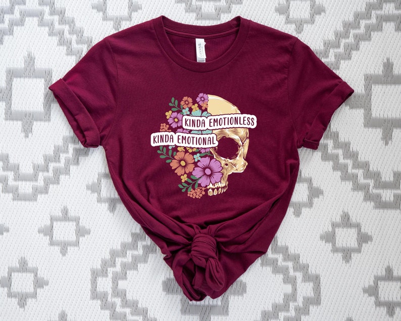 Puede incluir: Una camiseta color burdeos con un gr&aacute;fico de calavera y un dise&ntilde;o floral. El texto "Kinda Emotionless Kinda Emotional" est&aacute; impreso en la camiseta.