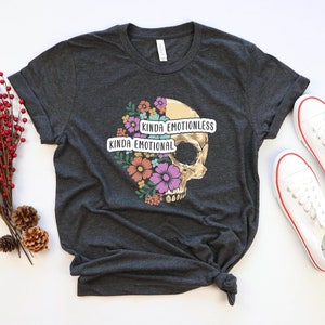 Puede incluir: Camiseta gris oscuro con un gr&aacute;fico de calavera y dise&ntilde;o floral. La calavera est&aacute; dividida en dos mitades con el texto "Kinda Emotional" en un lado y "Kinda Emotionless" en el otro.