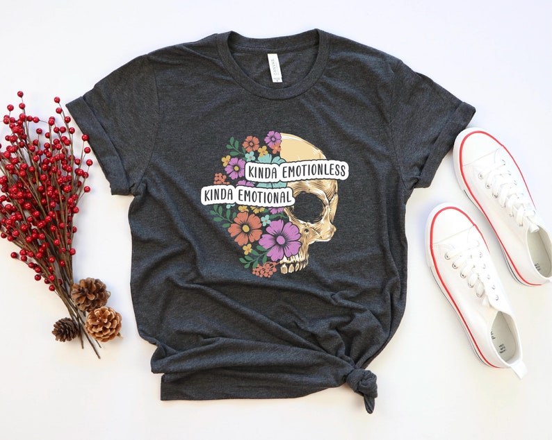 Puede incluir: Camiseta gris oscuro con un gr&aacute;fico de calavera y dise&ntilde;o floral. La calavera est&aacute; dividida en dos mitades con el texto "Kinda Emotional" en un lado y "Kinda Emotionless" en el otro.