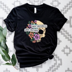 Puede incluir: Camiseta negra con un gr&aacute;fico de calavera rodeado de flores. El texto "Kinda Emotionless Kinda Emotional" est&aacute; impreso en la camiseta.