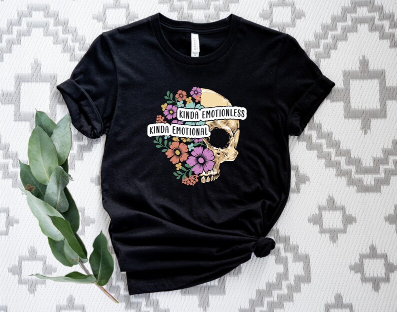 Puede incluir: Camiseta negra con un gr&aacute;fico de calavera rodeado de flores. El texto "Kinda Emotionless Kinda Emotional" est&aacute; impreso en la camiseta.