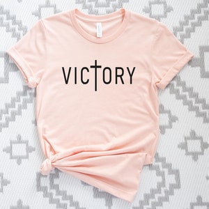 K&ouml;nnte beinhalten: Ein hellrosa T-Shirt mit einem schwarzen Kreuz und dem Wort "VICTORY" auf der Vorderseite.
