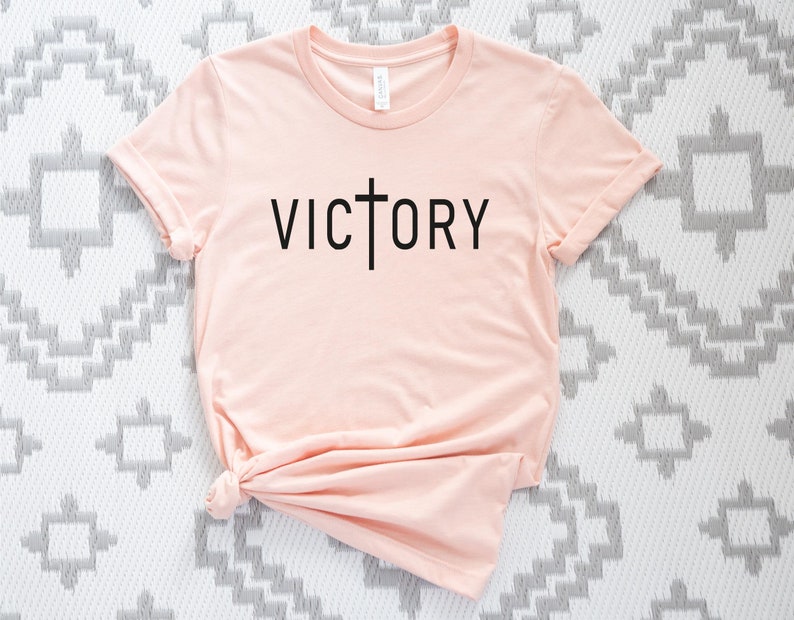 K&ouml;nnte beinhalten: Ein hellrosa T-Shirt mit einem schwarzen Kreuz und dem Wort "VICTORY" auf der Vorderseite.