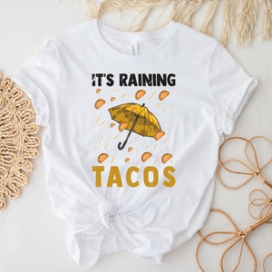 Peut inclure: T-shirt blanc avec un parapluie jaune et le texte "Il pleut des tacos" imprim&eacute; dessus.