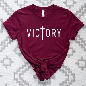 K&ouml;nnte beinhalten: Ein maroonfarbenes T-Shirt mit einem wei&szlig;en Kreuz und dem Wort "VICTORY" auf der Vorderseite.