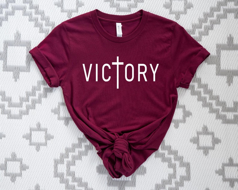 K&ouml;nnte beinhalten: Ein maroonfarbenes T-Shirt mit einem wei&szlig;en Kreuz und dem Wort "VICTORY" auf der Vorderseite.