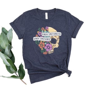 Puede incluir: Una camiseta azul oscuro con un gr&aacute;fico de calavera rodeado de flores coloridas. La calavera tiene una pancarta que dice "Kinda Emotionless" y otra pancarta que dice "Kinda Emotional".