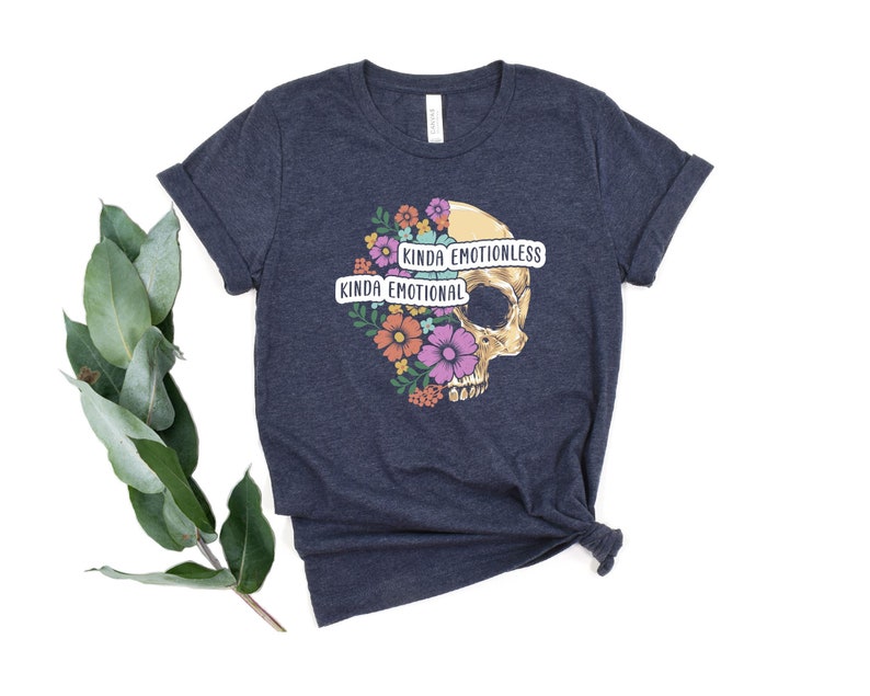Puede incluir: Una camiseta azul oscuro con un gr&aacute;fico de calavera rodeado de flores coloridas. La calavera tiene una pancarta que dice "Kinda Emotionless" y otra pancarta que dice "Kinda Emotional".