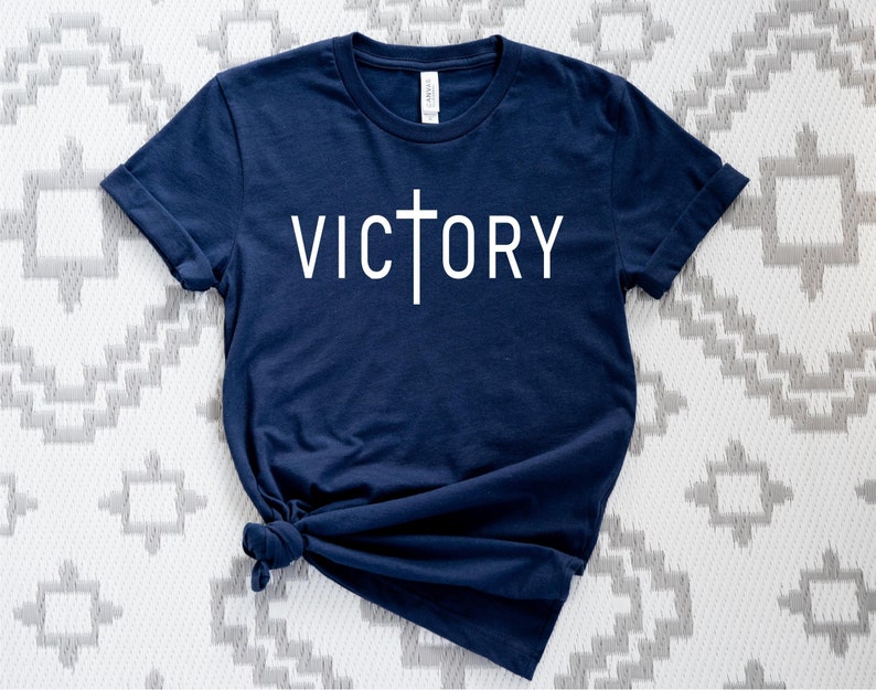 K&ouml;nnte beinhalten: Ein marineblaues T-Shirt mit einem wei&szlig;en Kreuz und dem Wort "VICTORY" auf der Vorderseite.