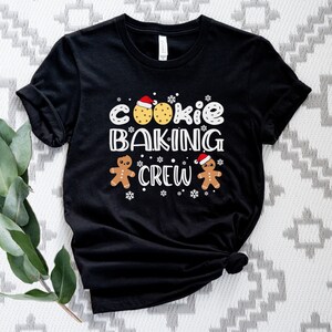 May include: Black t-shirt with a white graphic that says "Cookie Baking Crew" with gingerbread men and cookies wearing Santa hats.