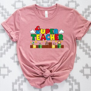 Puede incluir: Una camiseta rosa con un gr&aacute;fico de un personaje de videojuego y el texto "SUPER TEACHER" en letras de colores.