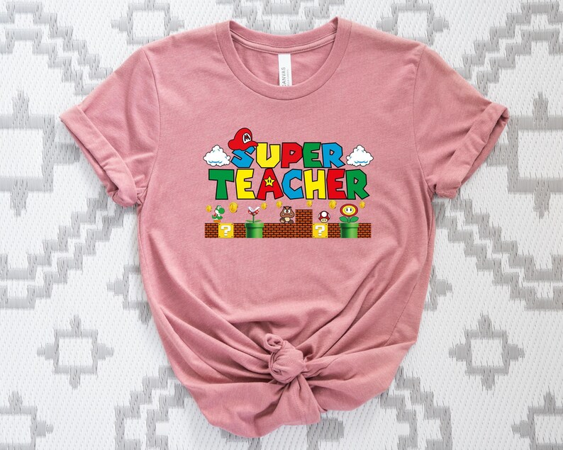 Puede incluir: Una camiseta rosa con un gr&aacute;fico de un personaje de videojuego y el texto "SUPER TEACHER" en letras de colores.