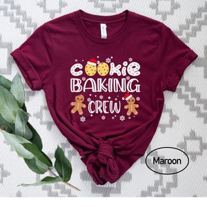May include: A maroon t-shirt with the text "Cookie Baking Crew" in white. The design includes two gingerbread men wearing Santa hats and a cookie with chocolate chips.