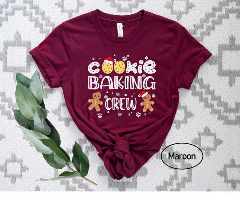 May include: A maroon t-shirt with the text "Cookie Baking Crew" in white. The design includes two gingerbread men wearing Santa hats and a cookie with chocolate chips.