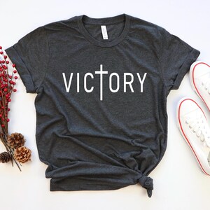 K&ouml;nnte beinhalten: Ein dunkelgraues T-Shirt mit einem wei&szlig;en Kreuz und dem Wort "VICTORY" auf der Vorderseite.