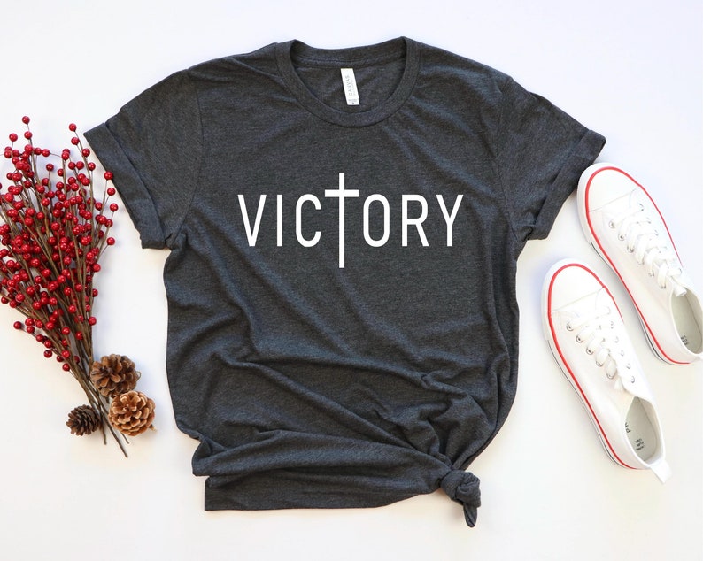 K&ouml;nnte beinhalten: Ein dunkelgraues T-Shirt mit einem wei&szlig;en Kreuz und dem Wort "VICTORY" auf der Vorderseite.