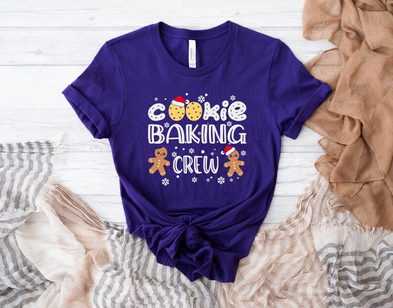 May include: Purple short-sleeved t-shirt with the text "Cookie Baking Crew" in white. The design includes two gingerbread men with red hats and a cookie with red icing.
