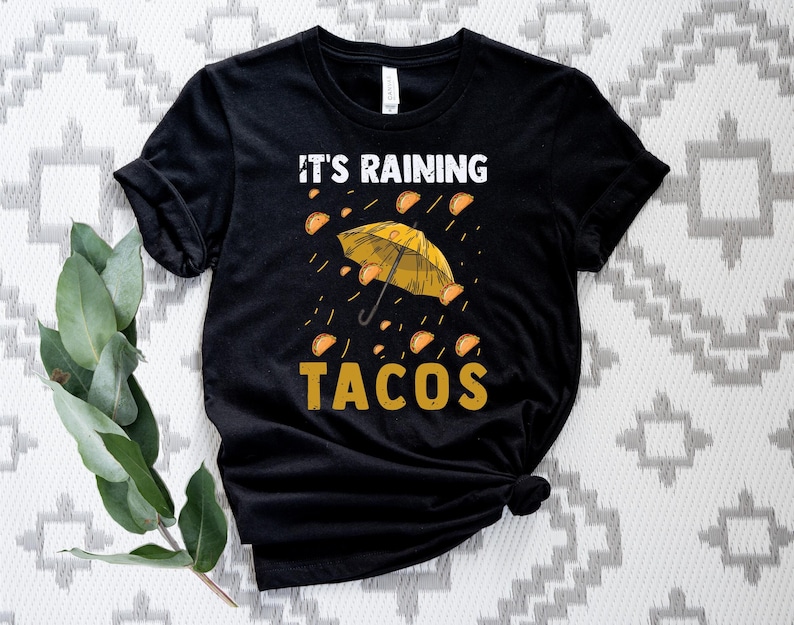 Peut inclure: T-shirt noir avec un graphique de tacos jaunes qui pleuvent du ciel avec un parapluie jaune. Le texte "It's Raining Tacos" est imprim&eacute; sur le t-shirt.