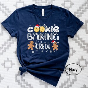 May include: A navy blue t-shirt with the text "Cookie Baking Crew" in white. The design includes two gingerbread men wearing Santa hats and two cookies with red frosting.