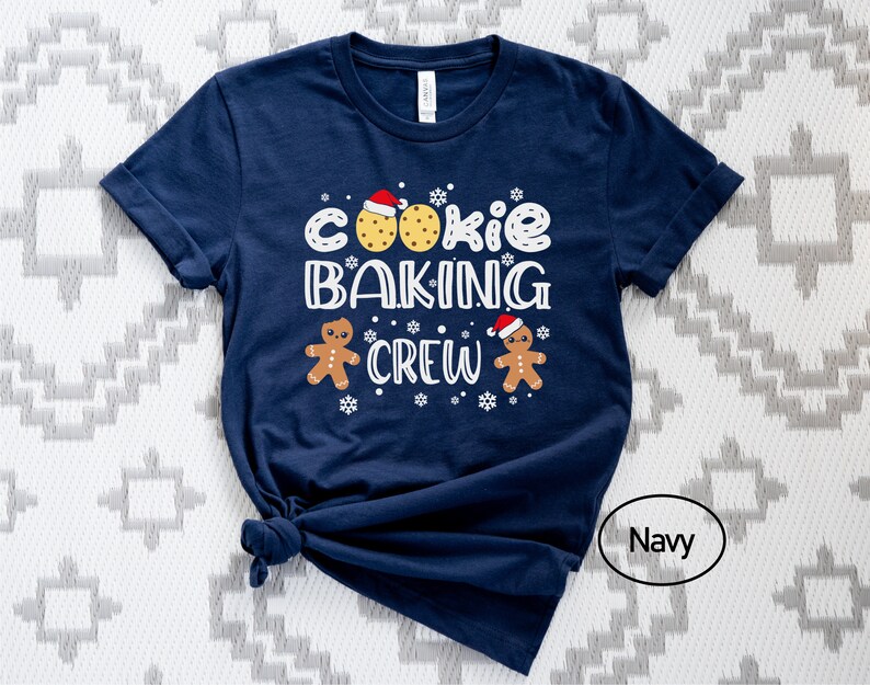 May include: A navy blue t-shirt with the text "Cookie Baking Crew" in white. The design includes two gingerbread men wearing Santa hats and two cookies with red frosting.